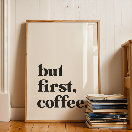 A framed poster with the text "but first, coffee" is displayed on a wooden floor next to a stack of books.