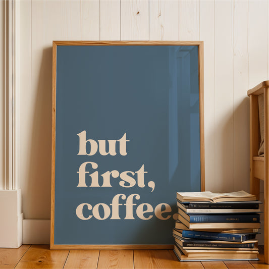 A framed poster with the text "but first, coffee" is displayed on a wooden floor next to a stack of books.