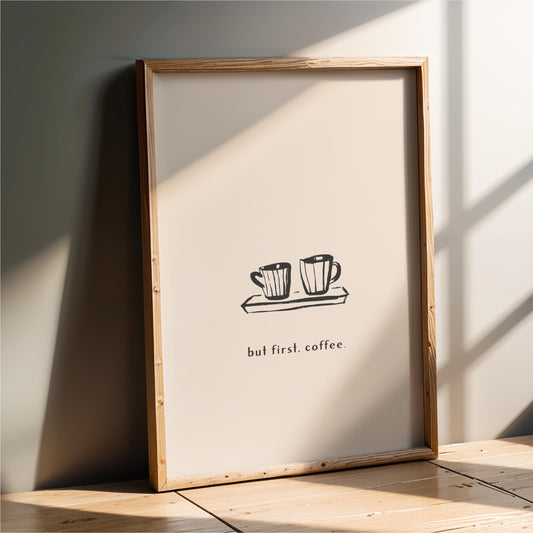 A framed poster with a simple black and white illustration of two coffee cups on a tray, accompanied by the text "but first. coffee."
