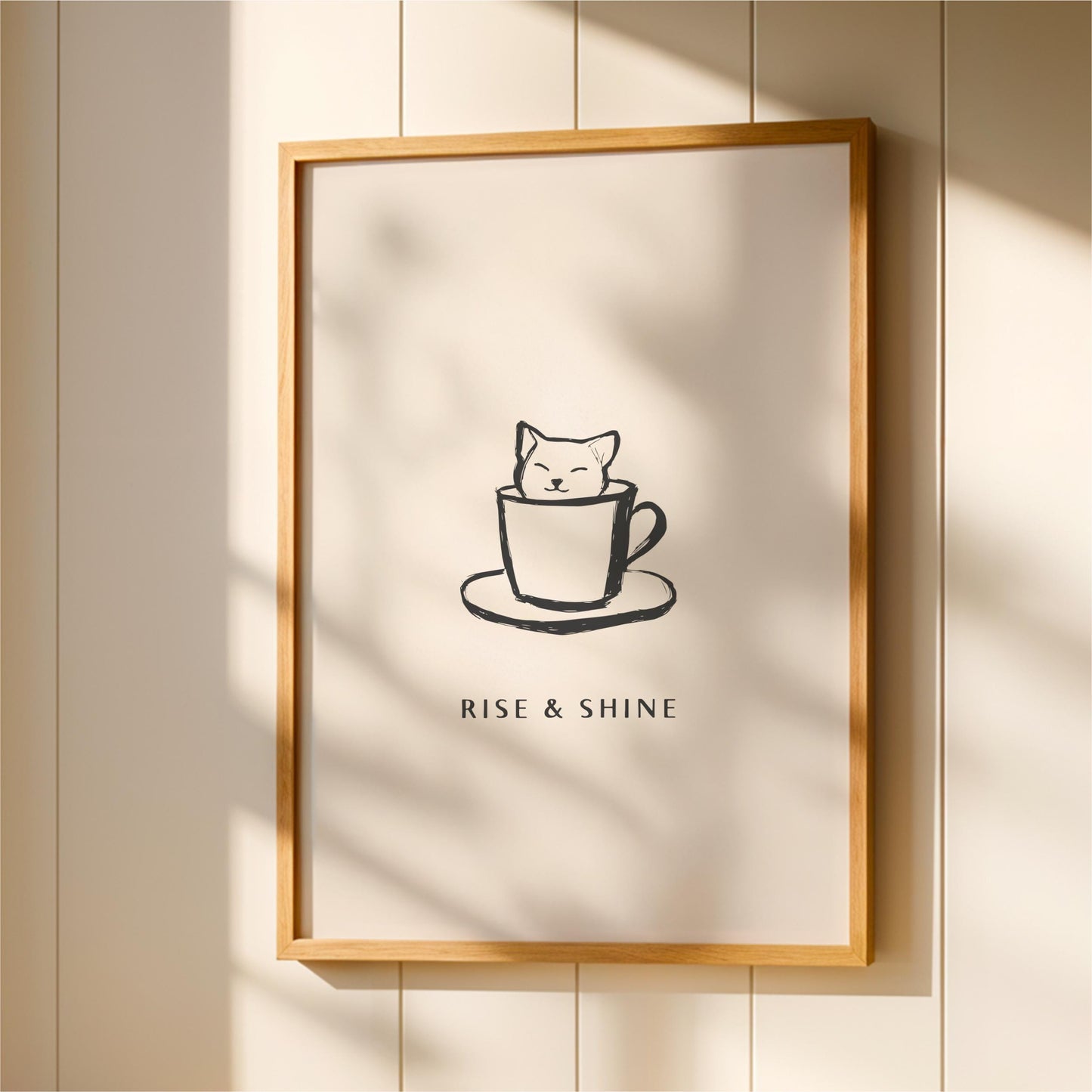 A framed poster with a simple black and white illustration of a cat sitting in a cup, accompanied by the text "RISE & SHINE" below the image.