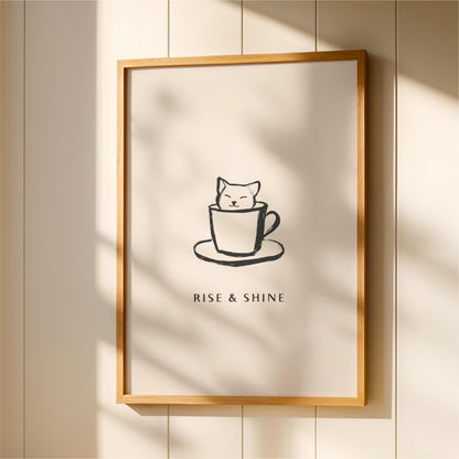 A framed poster with a simple black and white illustration of a cat sitting in a cup, accompanied by the text "RISE & SHINE" below the image.