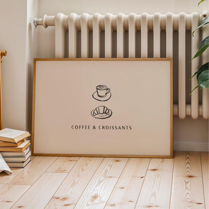 A framed poster with the text "COFFEE & CROISSANTS" and two illustrations of a coffee cup and a croissant, placed on a wooden floor next to a radiator.