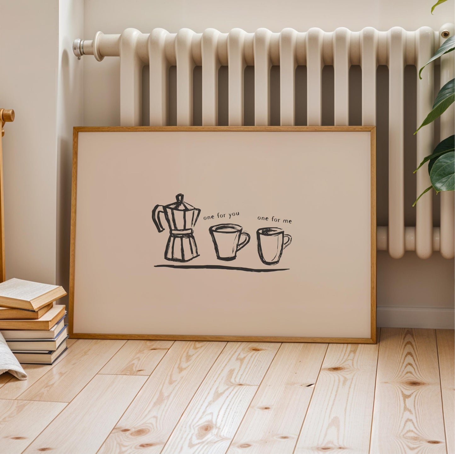A framed print with a simple black and white illustration of a coffee maker, two mugs, and text that reads "one for me" is displayed on a wooden floor next to a radiator.