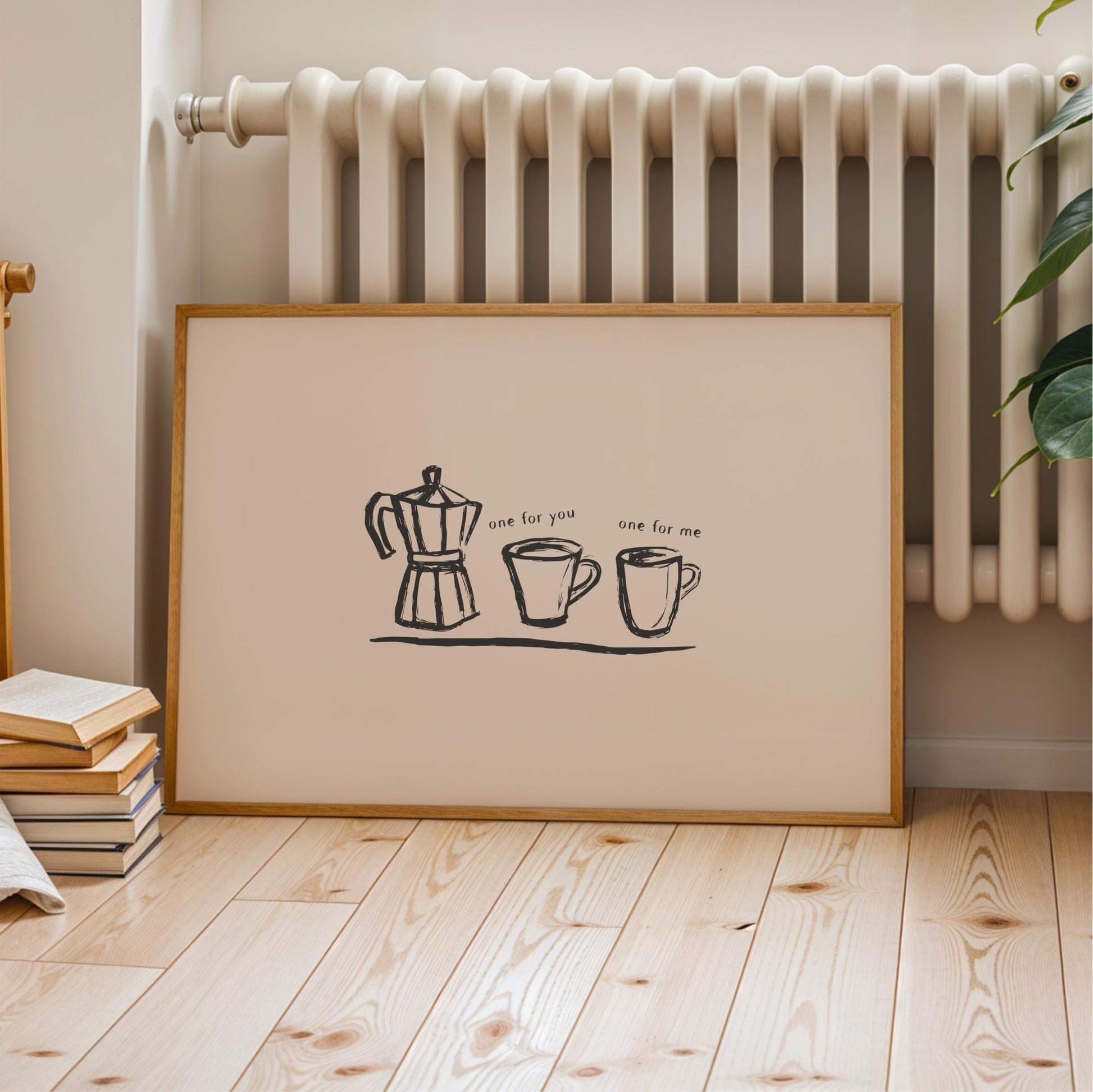 A framed print with a simple black and white illustration of a coffee maker, two mugs, and text that reads "one for me" is displayed on a wooden floor next to a radiator.