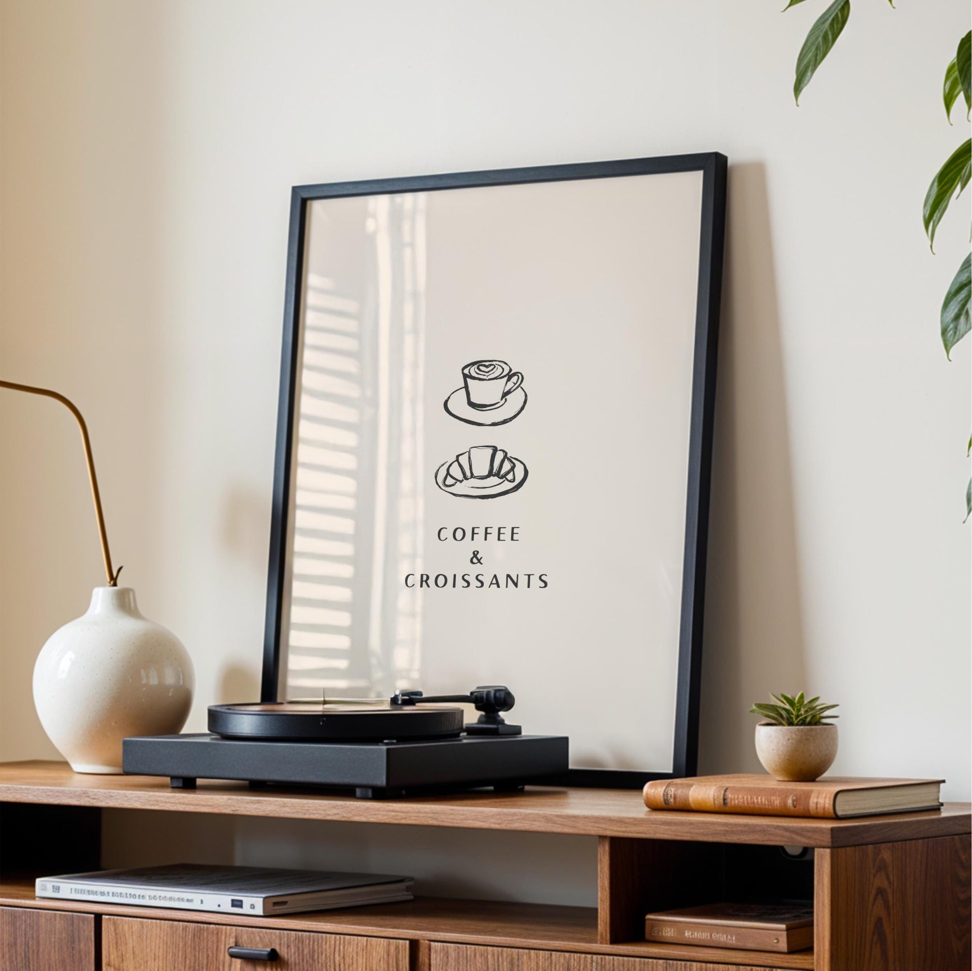 a framed poster with the text "COFFEE & CROISSANTS" on it, placed on a wooden shelf next to a white vase and a small plant.