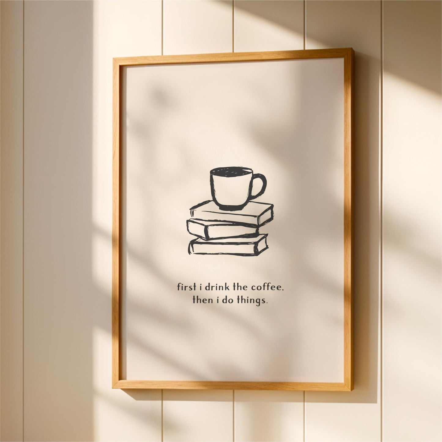 A framed poster with a simple black and white illustration of a cup of coffee on top of a stack of books, accompanied by the text "first I drink the coffee. Then I do things."