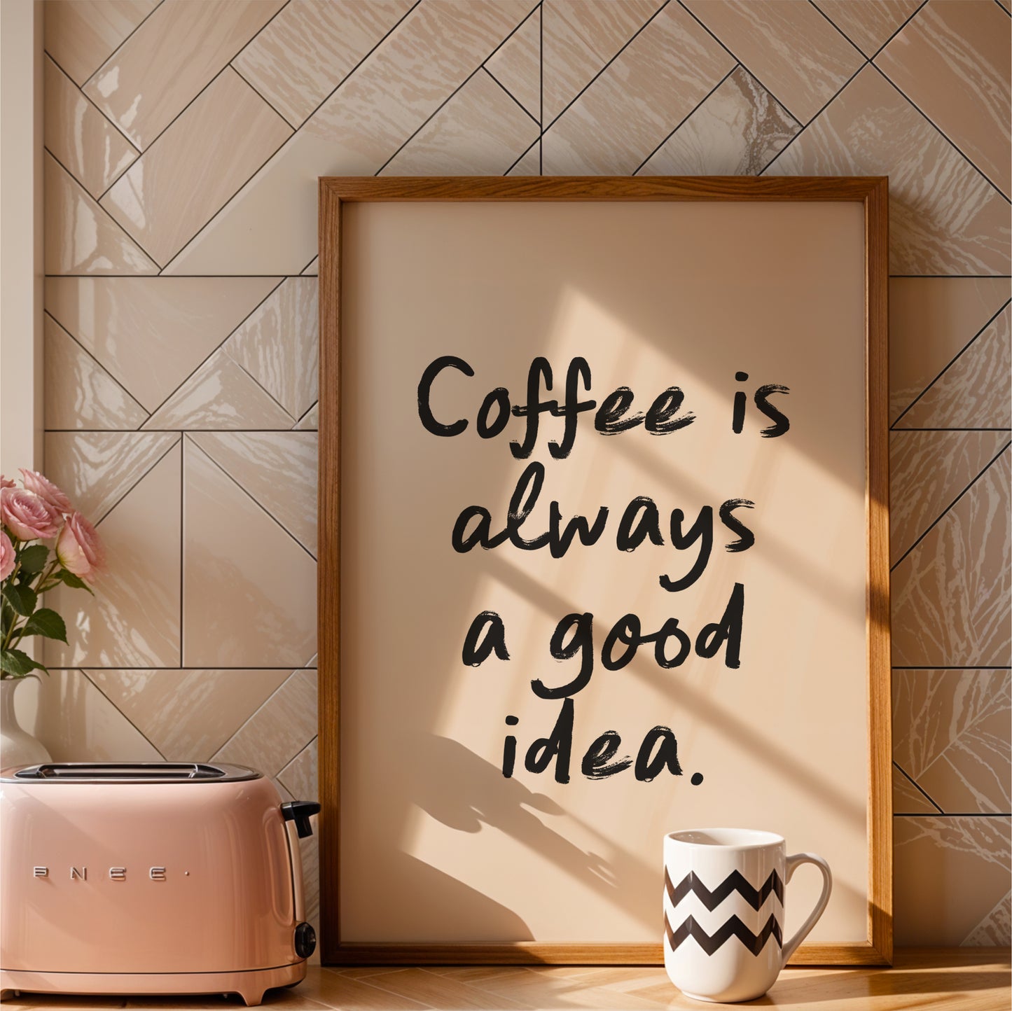 a framed quote that reads "Coffee is always a good idea." The quote is written in a casual, handwritten style. The frame is placed next to a pink toaster, and there is a white mug with a zigzag pattern on it. The background has a tiled wall, and there is a vase with pink roses on the counter.