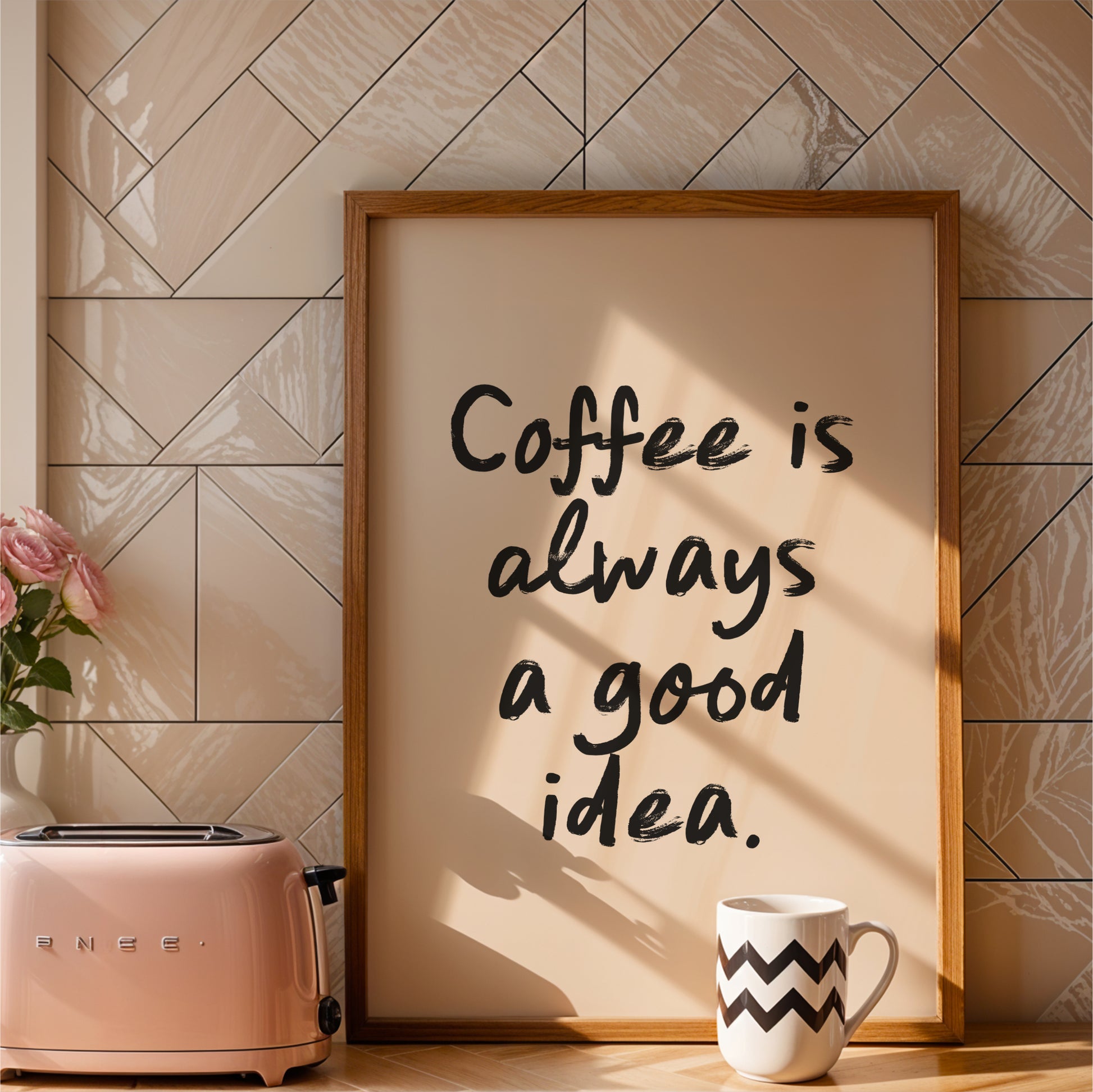 a framed quote that reads "Coffee is always a good idea." The quote is written in a casual, handwritten style. The frame is placed next to a pink toaster, and there is a white mug with a zigzag pattern on it. The background has a tiled wall, and there is a vase with pink roses on the counter.