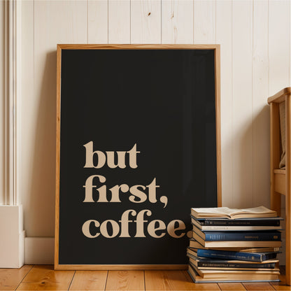 A framed poster with the text "but first, coffee" is displayed on a wooden floor next to a stack of books.