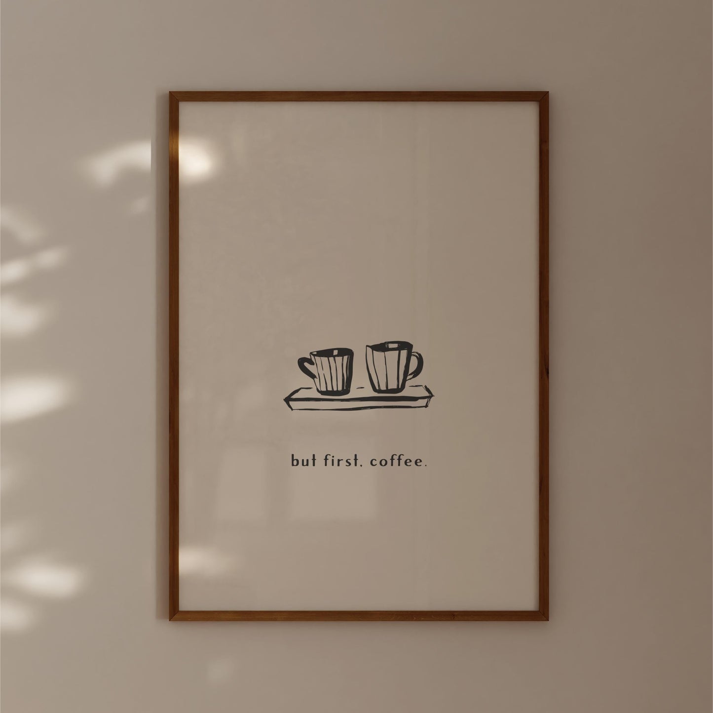 A framed print with a simple black and white illustration of two coffee cups on a tray, accompanied by the text "but first, coffee."