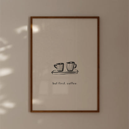A framed print with a simple black and white illustration of two coffee cups on a tray, accompanied by the text "but first, coffee."