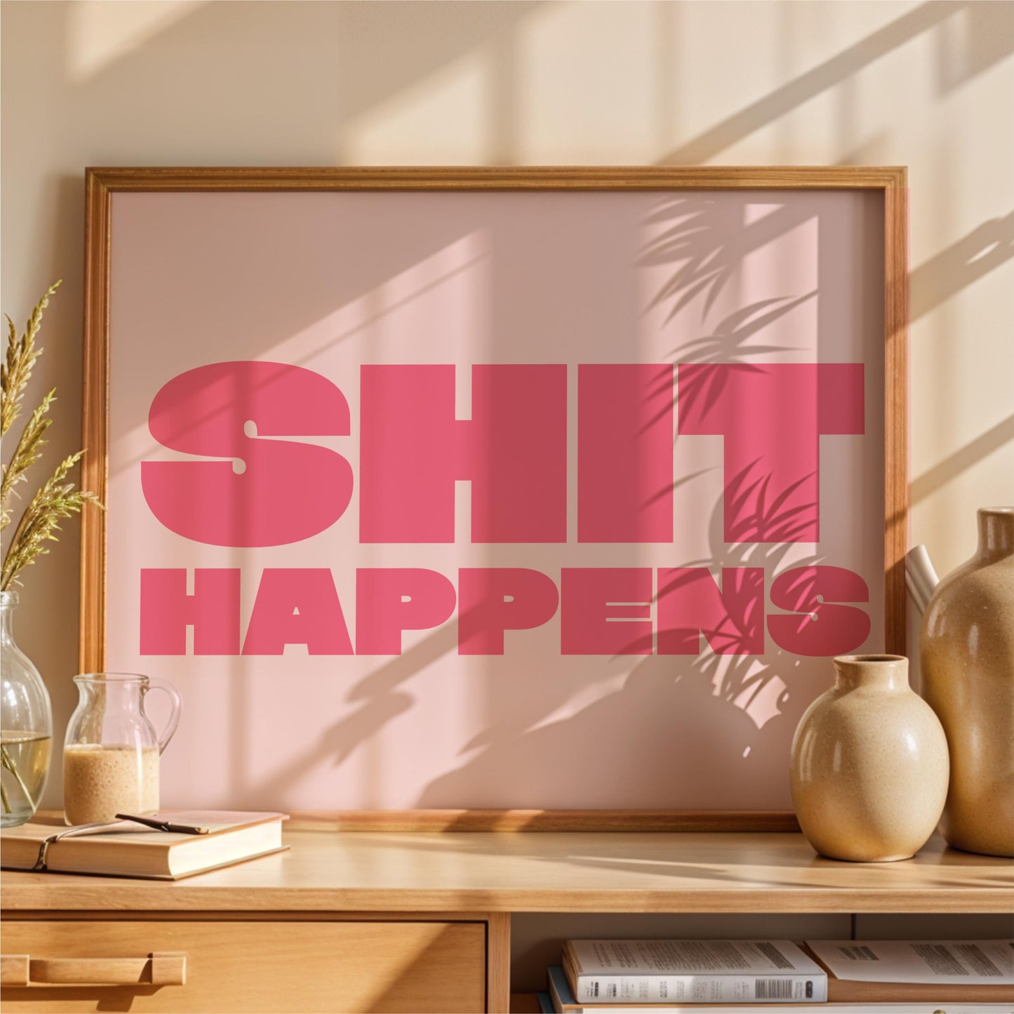 a framed poster with the text "SHIT HAPPENS" in large pink letters, placed on a wooden shelf next to a vase and some books.