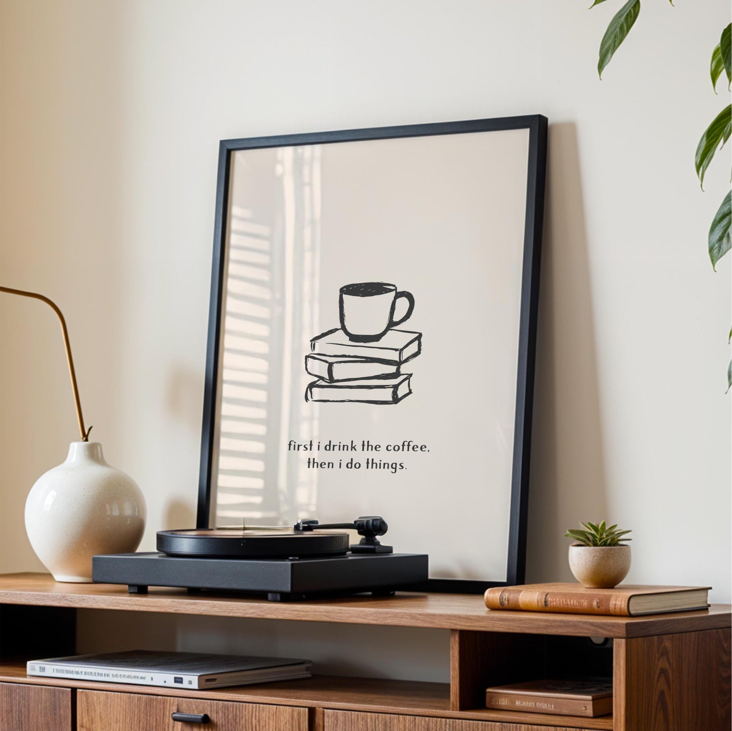 a framed poster with a simple black and white illustration of a coffee cup on a stack of books, accompanied by the text "first I drink the coffee. then I do things." The poster is displayed on a wooden shelf, with a white vase, a record player, and a stack of books nearby.
