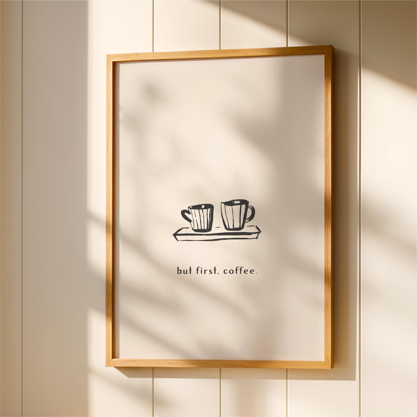 A framed print with a simple black and white illustration of two coffee cups on a tray, accompanied by the text "but first, coffee."