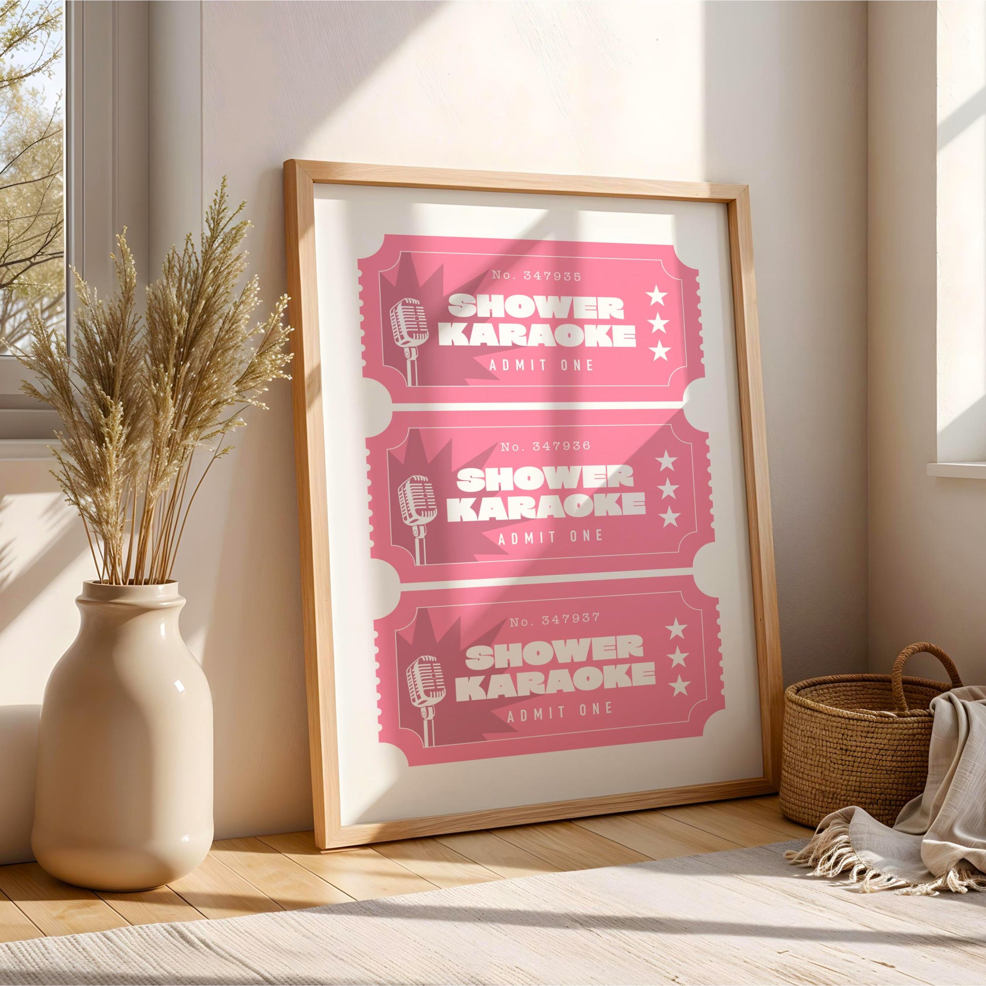 A framed poster advertising karaoke events, with three pink tickets featuring a microphone and the text "SHOWER KARAOKE" and "RUMIT ONE" printed on it. The poster is displayed on a wooden floor next to a vase with dried plants, and a woven basket is visible in the background.
