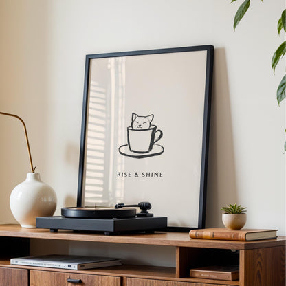 a framed poster with a cat illustration and the text "RISE & SHINE" on a wooden shelf.