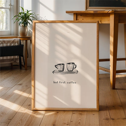 A framed poster with a simple black and white illustration of two coffee cups on a tray, accompanied by the text "but first coffee" in a casual, handwritten style.