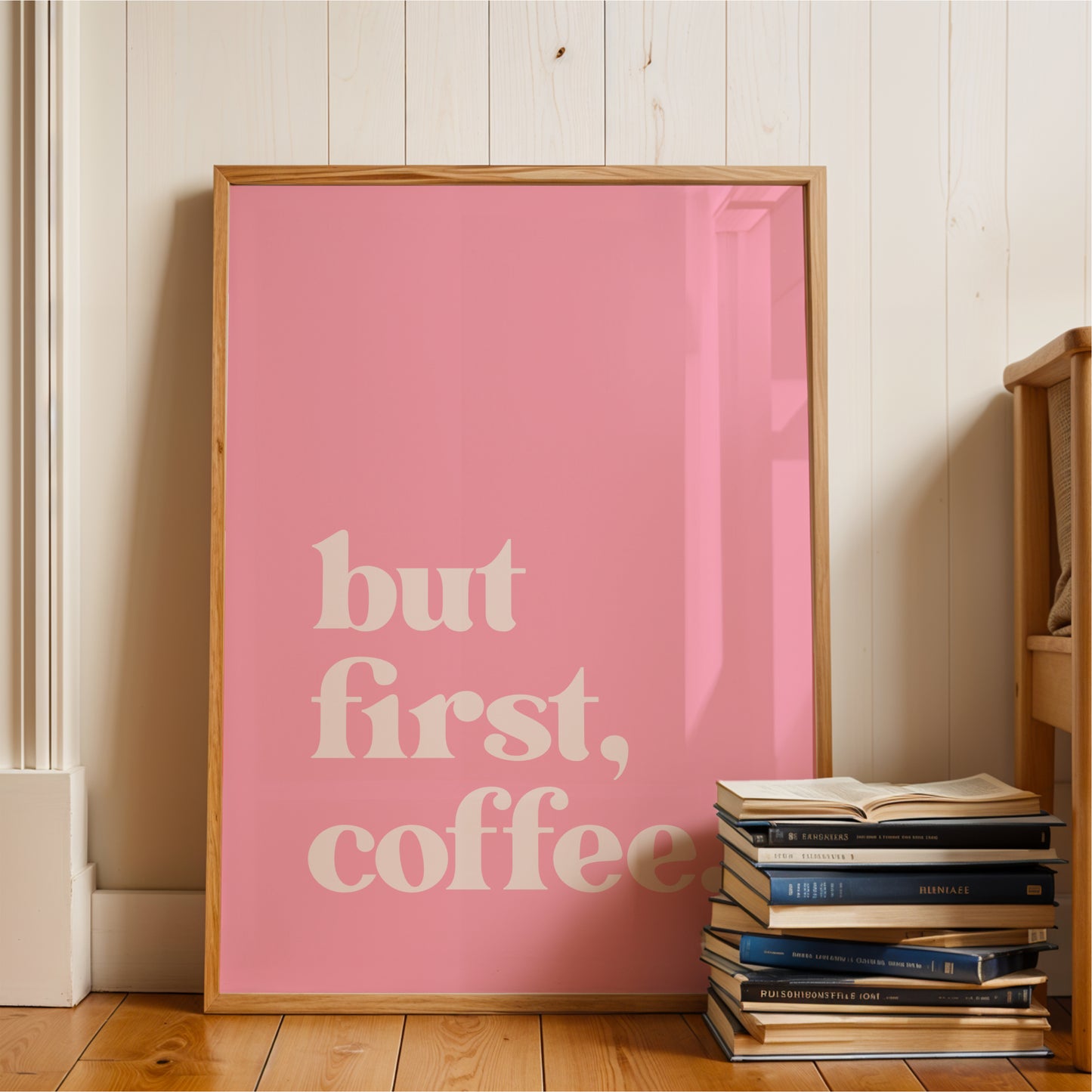 A pink poster with the text "but first, coffee" is displayed on a wooden floor next to a stack of books.