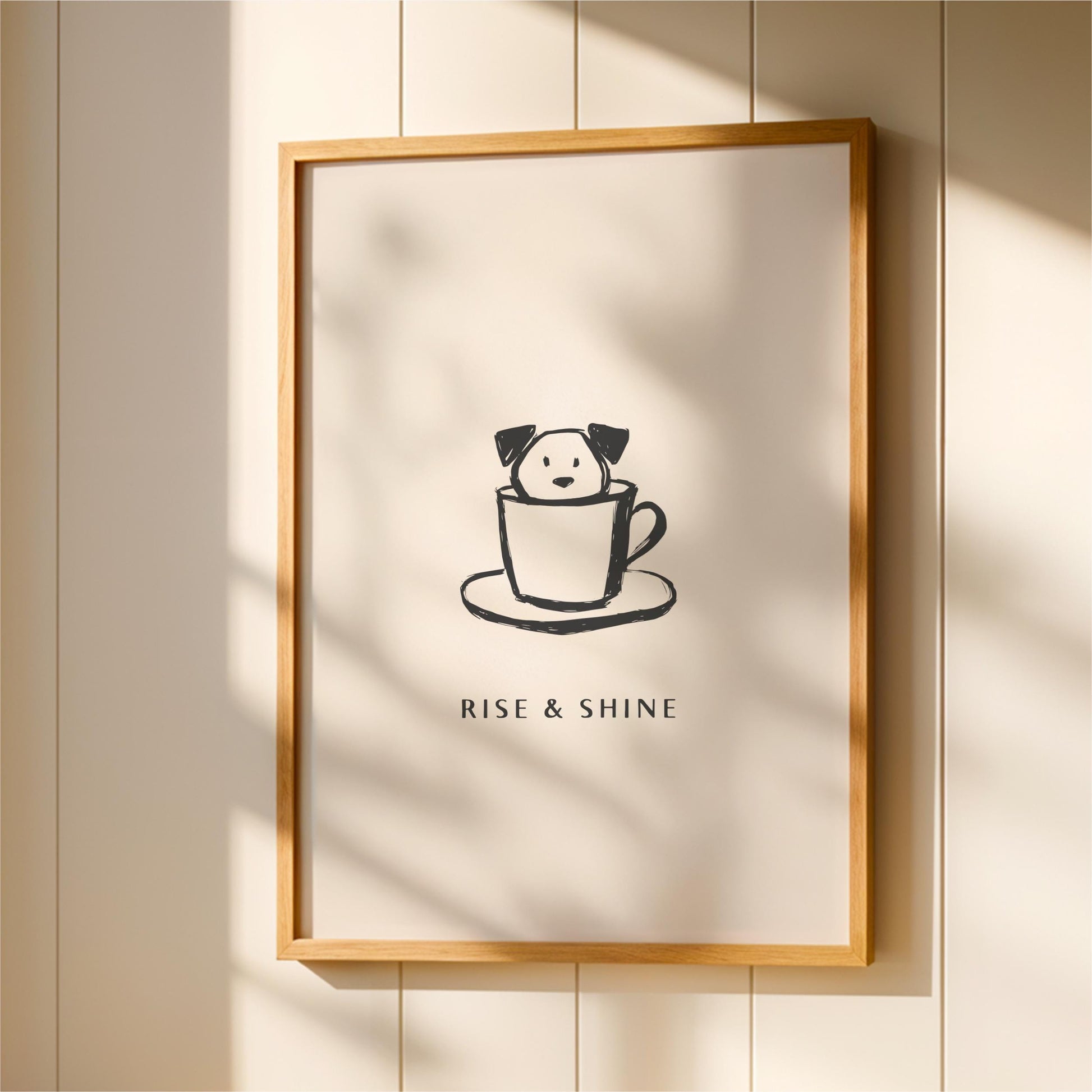 A framed poster with a black and white illustration of a dog in a cup, accompanied by the text "RISE & SHINE" below the image.