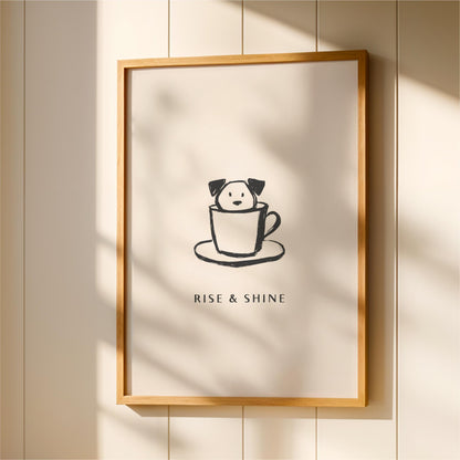 A framed poster with a black and white illustration of a dog in a cup, accompanied by the text "RISE & SHINE" below the image.