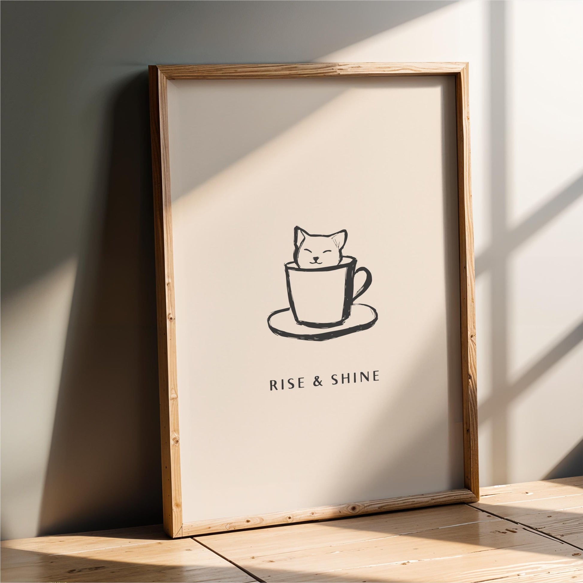 A framed poster with a simple black and white illustration of a cat sitting in a cup, accompanied by the text "RISE & SHINE" below the image.