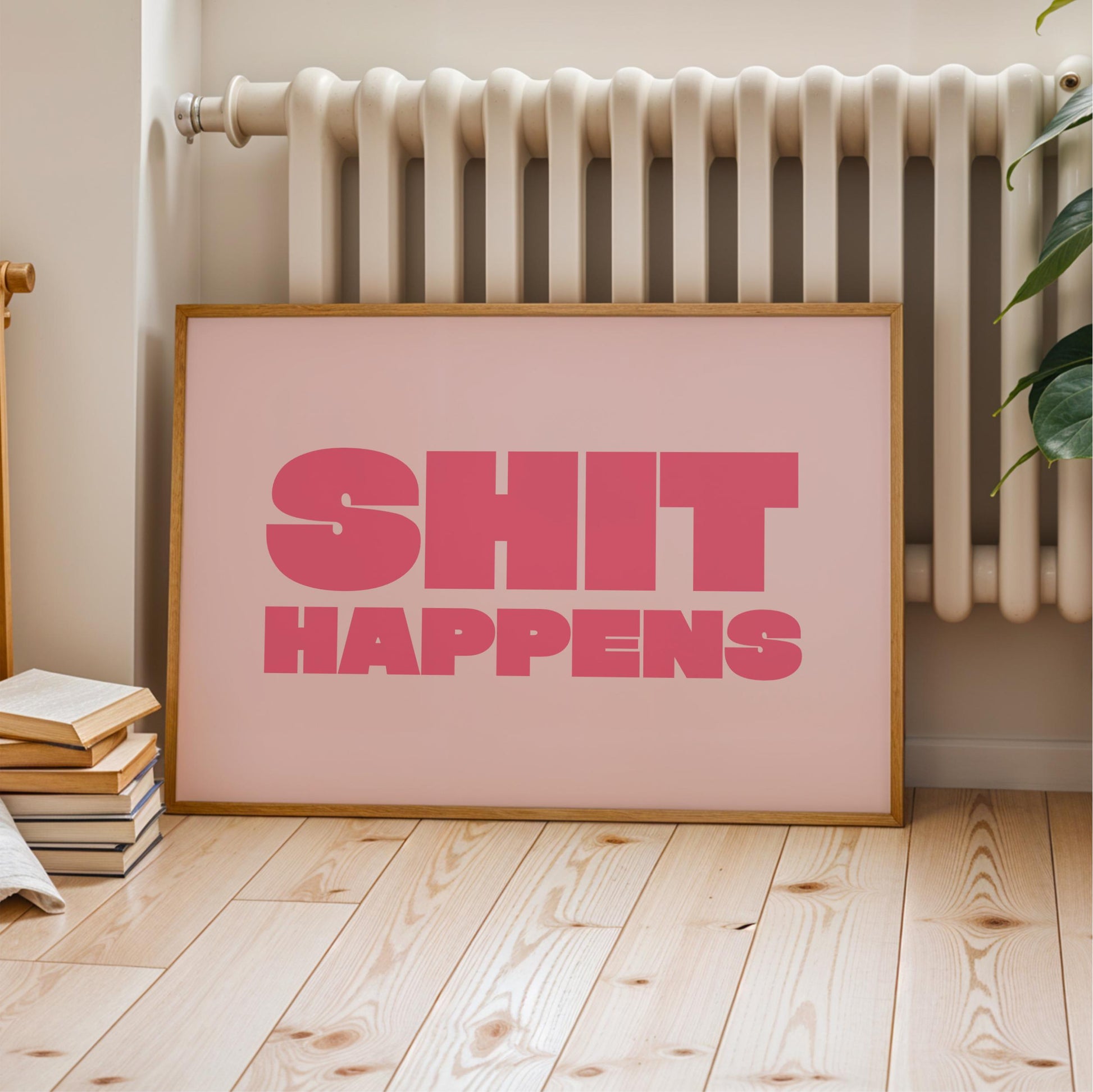 A framed poster with the text "SHIT HAPPENS" in pink letters is displayed on a wooden floor next to a radiator.