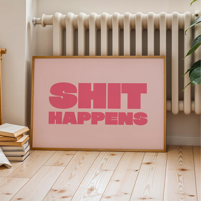 A framed poster with the text "SHIT HAPPENS" in pink letters is displayed on a wooden floor next to a radiator.