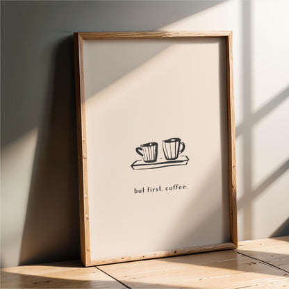 A framed poster with a simple black and white illustration of two coffee cups on a tray, accompanied by the text "but first. coffee."