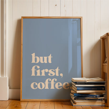A framed poster with the text "but first, coffee" is displayed on a wooden floor next to a stack of books.