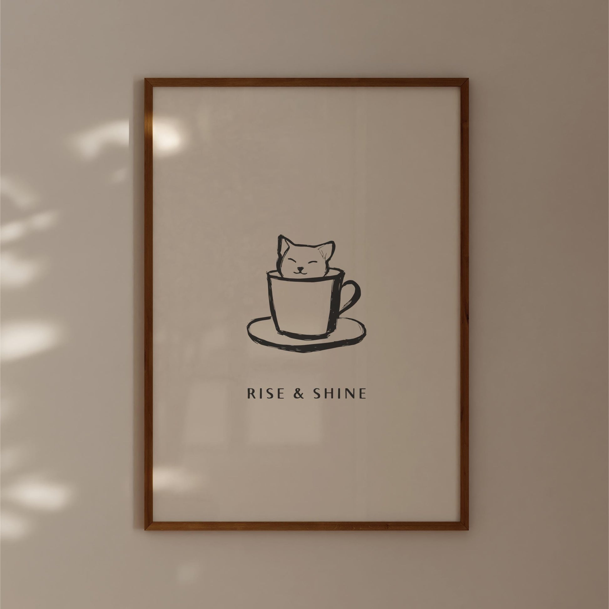 A framed poster with a simple black and white illustration of a cat sitting inside a cup, accompanied by the text "RISE & SHINE" below the image.