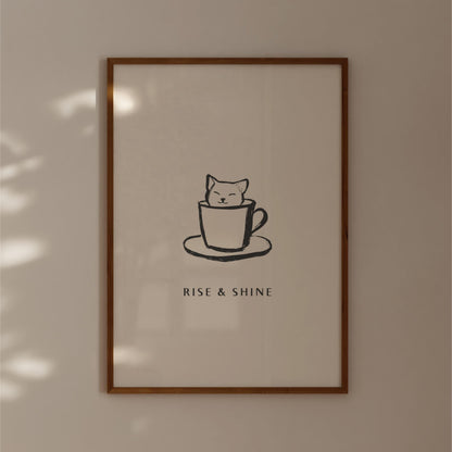 A framed poster with a simple black and white illustration of a cat sitting inside a cup, accompanied by the text "RISE & SHINE" below the image.