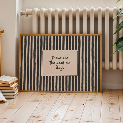 A framed poster with a black and white striped pattern and the text "These are the good old days" is displayed on a wooden floor next to a white radiator.