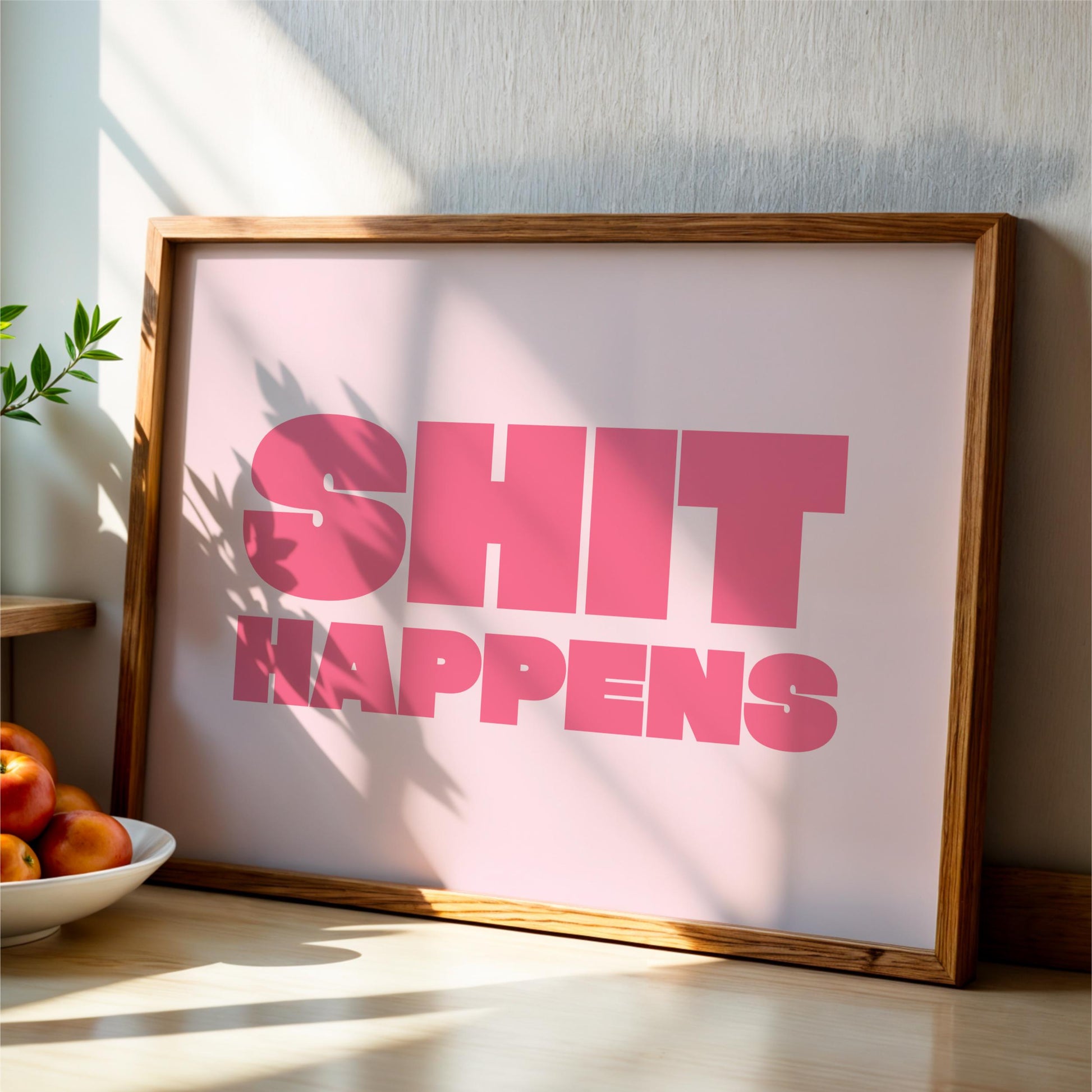 A framed poster with the text "SHIT HAPPENS" in large pink letters, placed on a wooden surface next to a bowl of fruit.