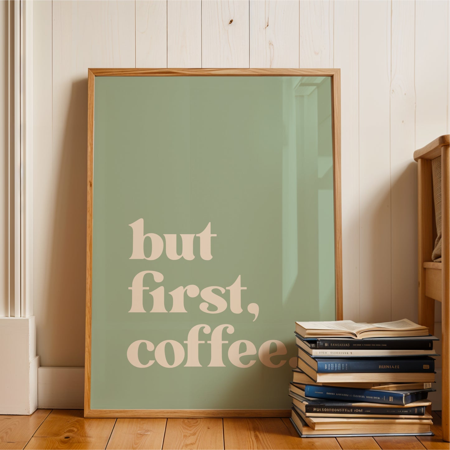 A framed poster with the text "but first, coffee" is displayed on a wooden floor next to a stack of books.