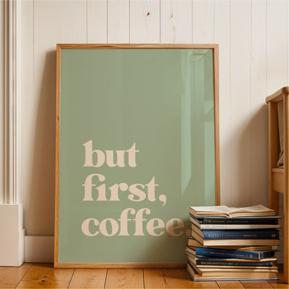 A framed poster with the text "but first, coffee" is displayed on a wooden floor next to a stack of books.