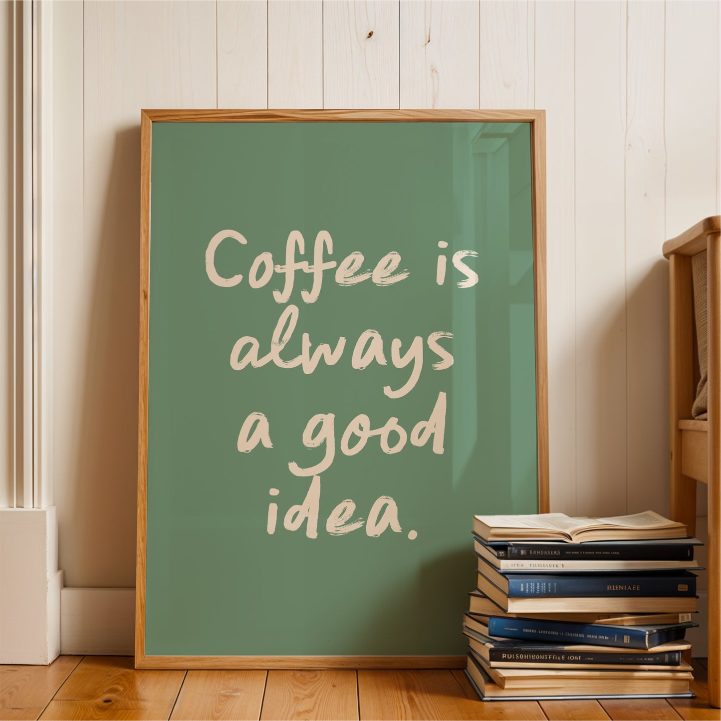 A framed poster with the text "Coffee is always a good idea" is displayed on a wooden floor next to a stack of books.